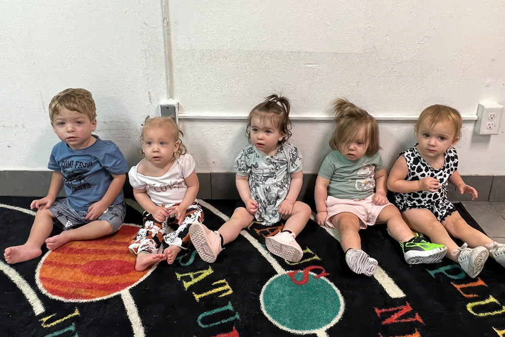 Five charming toddlers sit in a row on a colorful rug displaying their curious gazes highlights the innocence.